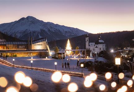 Un paesaggio invernale pittoresco con montagne innevate e un'illuminazione festiva dell'albero di Natale. Sullo sfondo si vede una chiesa e un edificio moderno.