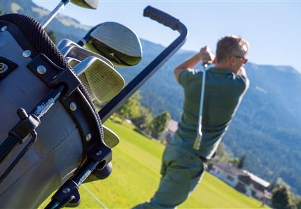 Ein Golfer schlägt einen Ball auf einem schönen Golfplatz mit Bergen im Hintergrund. Im Vordergrund steht eine Golftasche.