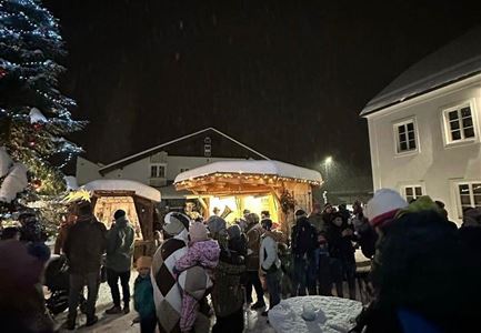 Eine Menschenmenge versammelt sich bei einem festlichen Stand in einer winterlichen Nacht. Die Umgebung ist schneebedeckt, und ein Weihnachtsbaum leuchtet im Hintergrund.