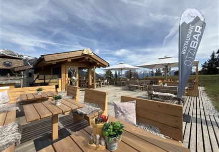 A cozy outdoor area of a restaurant with wooden tables and chairs. In the background, mountains and a blue sky can be seen.