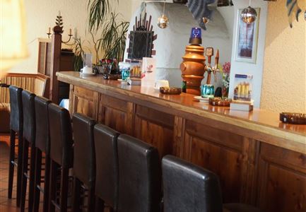 Eine rustikale Bar mit dunklem Holz und schwarzen Barhockern. Auf der Theke stehen verschiedene Getränke und Dekorationselemente.