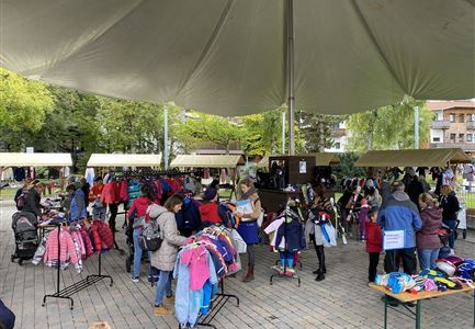 Een levendige markt met mensen die kinder kleding en accessoires bekijken. Kramen staan onder een groot tentzeil en zijn omringd door bomen.