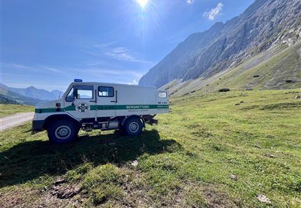 Ein Bergrettungsfahrzeug steht auf einer grünen Wiese in den Alpen. Im Hintergrund sind majestätische Berge und ein klarer blauer Himmel zu sehen.