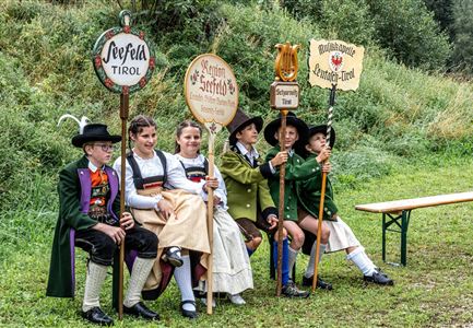 Eine Gruppe von Kindern in traditionellen Trachten sitzt auf einer Bank im Freien. Sie halten Schilder mit verschiedenen Aufschriften und blicken freundlich in die Kamera.