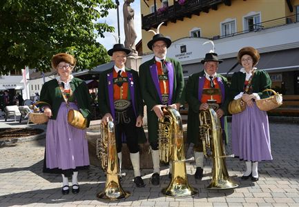 Five musicians in traditional attire stand in a sunny city. They hold large tubas and smile at the camera.