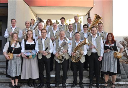 Eine große Musikkapelle steht vor einer Bergkulisse. Die Mitglieder tragen traditionelle Kleidung und halten Instrumente in der Hand.