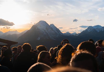 A large crowd stands in front of majestic mountains at sunset. The sky is partially cloudy and the atmosphere seems festive.