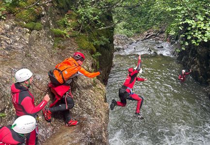Eine Gruppe von Menschen in Neoprenanzügen steht an einem Felsen und beobachtet, wie eine Person ins Wasser springt. Um sie herum ist eine grüne, bewaldete Umgebung.