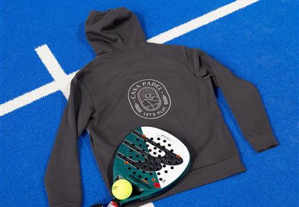 Ein graues Sweatshirt liegt auf einem blauen Sportplatz, neben einem Paddel und einem Tennisball. Das Sweatshirt zeigt ein Logo auf der Rückseite.
