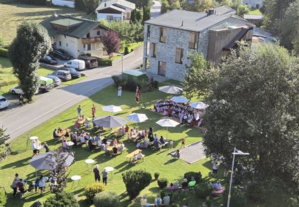 Eine belebte Versammlung im Freien mit vielen Menschen, die unter Sonnenschirmen sitzen. Im Hintergrund sind Gebäude und eine grüne Landschaft zu sehen.