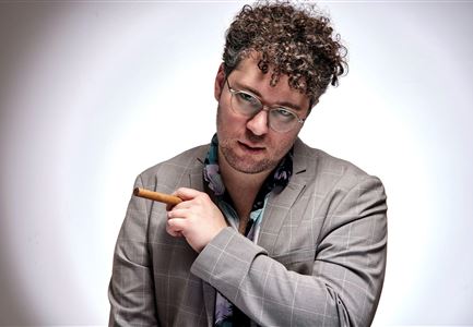 A stylish man in a gray suit with curly hair. He holds a cigar and looks directly at the camera.