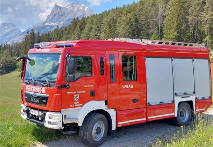 Ein rotes Feuerwehrfahrzeug steht auf einem Weg in einer Landschaft mit Bergen und Bäumen. Die Umgebung ist grün und sonnig.