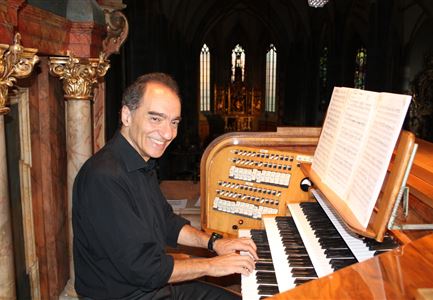 Ein Mann sitzt an einer Orgel und spielt. Im Hintergrund sieht man eine beeindruckende Kirche mit hohen Fenstern.