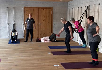 Eine Gruppe von Personen macht Fitnessübungen in einem modernen Raum. Der Boden ist aus Holz, und es gibt mehrere Yogamatten und Trainingsgeräte.