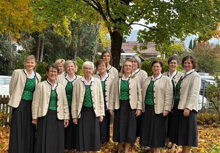 A group of women stands on a stage in traditional attire. The women wear green and black dresses and smile at the camera.