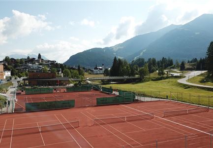 Ein Tennisplatz mit roter Asche in einer malerischen Landschaft. Im Hintergrund sind Berge und eine grüne Wiese zu sehen.