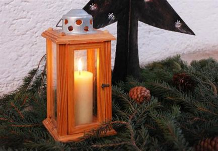 A rustic lantern with a burning candle stands on a bed of fir branches. In the background, a wooden Christmas tree can be seen.