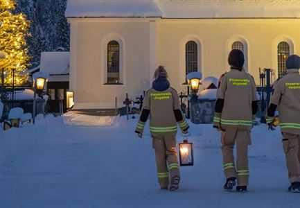 Drei Personen in Feuerwehranzügen gehen mit Laternen durch eine verschneite Landschaft. Im Hintergrund ist ein beleuchteter Baum neben einem Gebäude zu sehen.