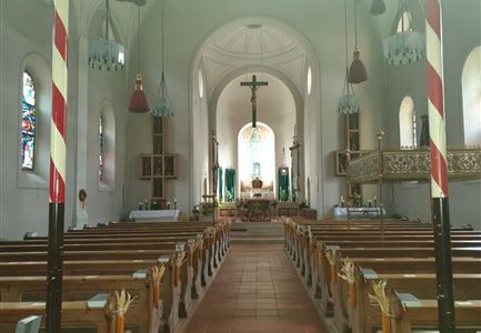 Ein großer Kirchenraum mit hölzernen Bänken und einer bunten Fensterfront. Im Hintergrund steht ein Altar mit einem Kreuz und weiteren dekorativen Elementen.