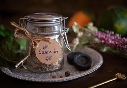 Ein Glas mit Kräutermischung und dem Etikett "Samhain" steht auf einem silbernen Tablett. Umgeben ist es von Trockenblumen und mehreren kleinen Deko-Elementen.