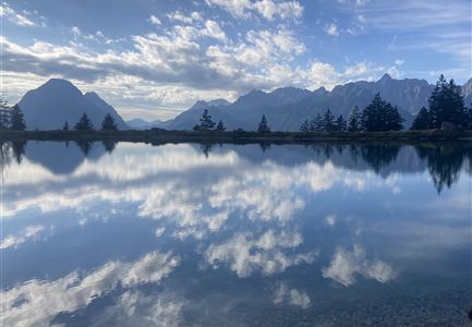 Ein ruhiger See spiegelt die umliegenden Berge und den wolkenlosen Himmel wider. Die Landschaft ist von grünen Bäumen umgeben.