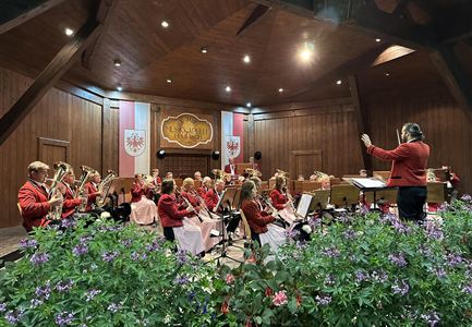 Eine Musikkapelle spielt in einem gemütlichen Saal. Im Vordergrund sind bunte Blumen zu sehen.