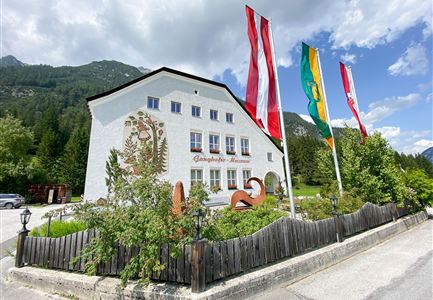 Ein traditionelles Gasthaus mit einer weißen Fassade und bunter Fahnen. Umgeben von grünen Bäumen und Bergen im Hintergrund.