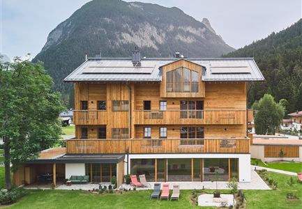 Ein modernes Holzgebäude mit mehreren Balkonen, umgeben von einer grünen Wiese. Im Hintergrund ist ein bewaldeter Berg zu sehen.