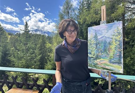 An artist stands next to her canvas and showcases a landscape painting. In the background, there are wooded mountains and a blue sky.
