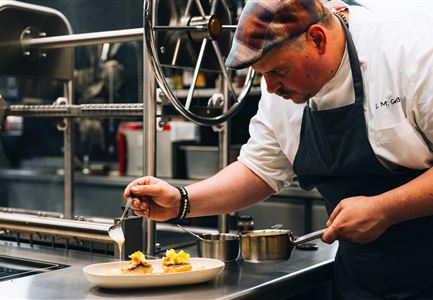 Ein Koch kreiert ein kunstvolles Gericht auf einem weißen Teller. Im Hintergrund sind Küchengeräte und eine moderne Küchenausstattung zu sehen.