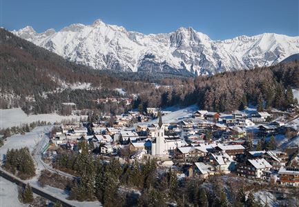Ein malerisches Dorf im Schnee, umgeben von hohen Bergen. Die Landschaft zeigt eine ruhige Winteridylle mit weißen Tälern und einer kleinen Kirche im Mittelpunkt.