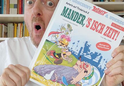 Ein Mann hält begeistert ein Comicbuch mit dem Titel "Mander, 's isch Zeit!" in der Hand. Vor ihm stehen kleine Figuren aus der Comicwelt.