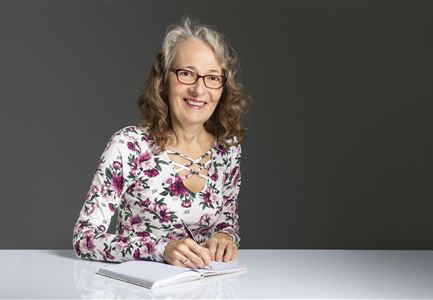 A smiling woman is sitting at a table and holding a pen in her hand. She is wearing a floral top and has curly, gray hair.