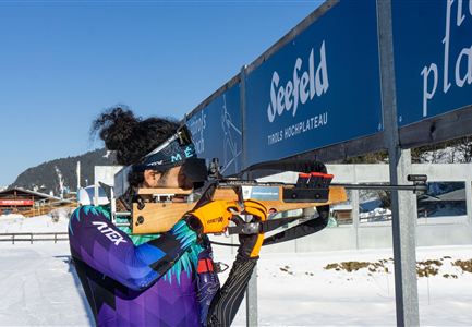 An athlete in a colorful outfit aims with a rifle at a target. In the background, snow-covered mountains and a sign reading "Seefeld" are visible.