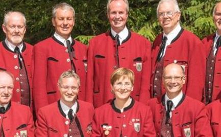 A group of twelve people in red jackets is standing outdoors. In the background, a green, wooded area can be seen.