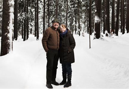 Ein Paar steht in einem verschneiten Wald. Hohe Bäume umgeben den Weg, und die Atmosphäre ist winterlich und ruhig.