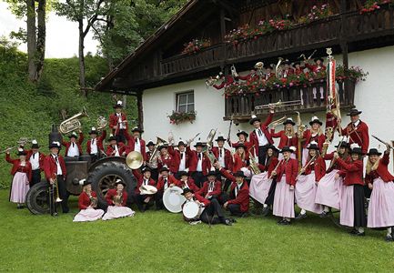 Una grande banda musicale in abbigliamento tradizionale si trova davanti a una casa colonica. Posano su un prato e tengono in mano vari strumenti musicali.