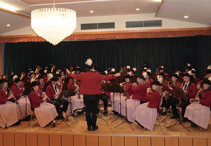 Ein großes Orchester spielt auf einer Bühne. Die Musiker tragen rote Jacken mit schwarzen Hüten und zeigen eine festliche Atmosphäre.