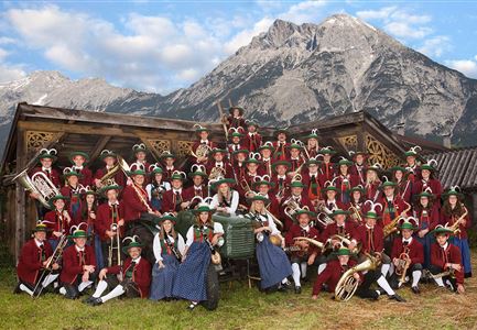 Eine große Musikkapelle steht vor einer Bergkulisse. Die Mitglieder tragen traditionelle Kleidung und halten Instrumente in der Hand.