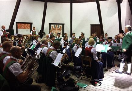 Eine große Musikkapelle steht vor einer Bergkulisse. Die Mitglieder tragen traditionelle Kleidung und halten Instrumente in der Hand.