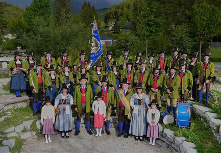 A group of people in traditional clothing gathers outdoors. They are smiling and holding their hands up.