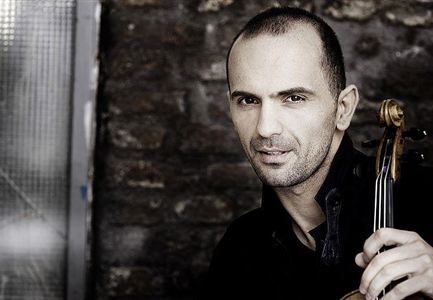 A man with a violin bow and a thoughtful expression stands in front of a stone wall. He is wearing a black shirt and has a short haircut.