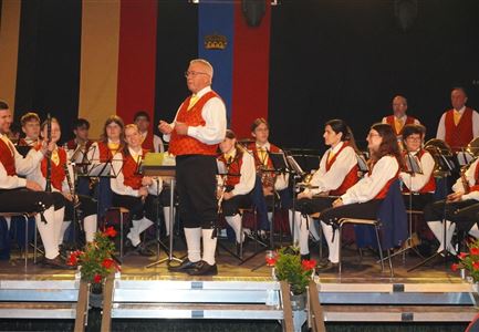 Eine Musikkapelle in traditioneller Kleidung spielt auf einer Bühne. Der Dirigent steht im Vordergrund und leitet das Orchester.