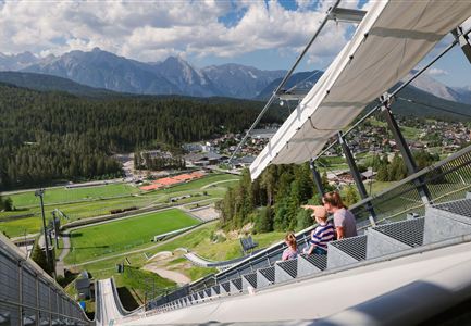 Eine sommerliche Landschaft mit einem aufregenden Steilhang und Blick auf die Berge. Im Vordergrund sitzen eine Familie auf einer Aussichtsplattform.