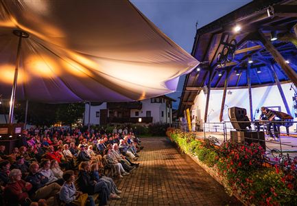 A crowd is sitting under a large sunshade and looking at the stage. The atmosphere is festive and there is beautiful lighting in the background.