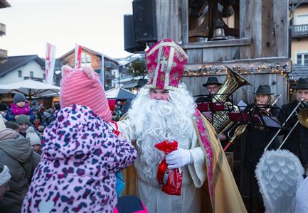 Ein festlich gekleideter Mann mit Bart hält Geschenke in der Hand. Im Hintergrund sind Menschen versammelt und eine Blaskapelle spielt Musik.