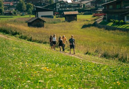 Eine Gruppe von fünf Personen geht auf einem Pfad durch eine grüne Wiese. Im Hintergrund sind Hütten und ein malerisches Dorf zu sehen.