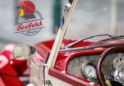 Ein klassisches Auto mit rotem Interieur und stilvollem Lenkrad. Im Hintergrund ist ein Logo mit einem Fahrerabzeichen zu sehen.