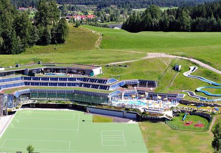 Un edificio alberghiero moderno in un ambiente verde. In primo piano si possono vedere campi da tennis e un'area piscina con scivoli.