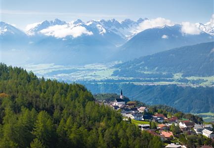 Eine malerische Landschaft mit Bergen und einem kleinen Dorf im Tal. Dichte Wälder umgeben die Siedlung, unter einem klaren Himmel.